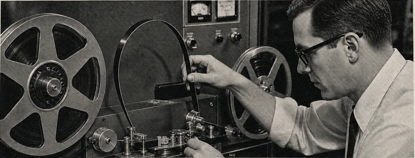 A technician threading a continuous loop of magnetic tape through a 1950s reel-to-reel machine.